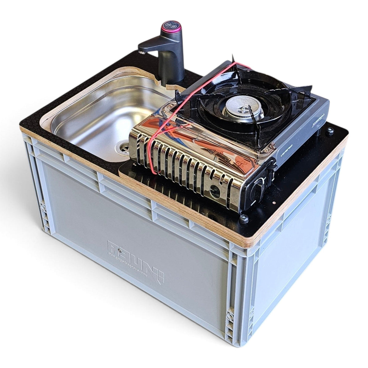 A compact outdoor kitchen setup mounted on a gray plastic storage crate. The unit includes a built-in stainless steel sink, a black countertop with a light wood trim, a portable gas stove with a built-in cooling fan, and a small black faucet with a pink accent.