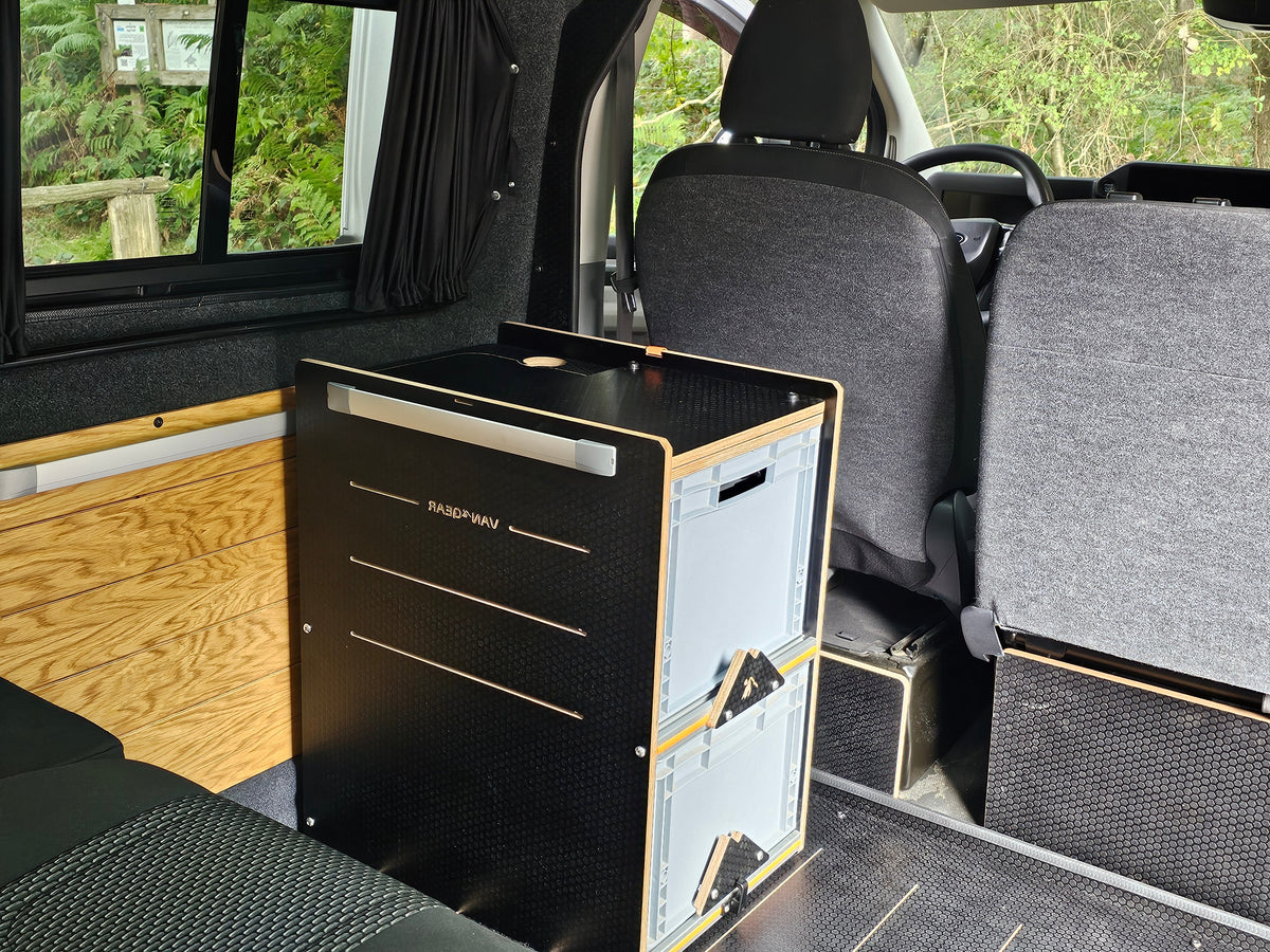 A black and wood Vaporesso travel system unit, likely a camp stove or kitchen cabinet, installed inside a vehicle's passenger compartment with gray seats and wood flooring.