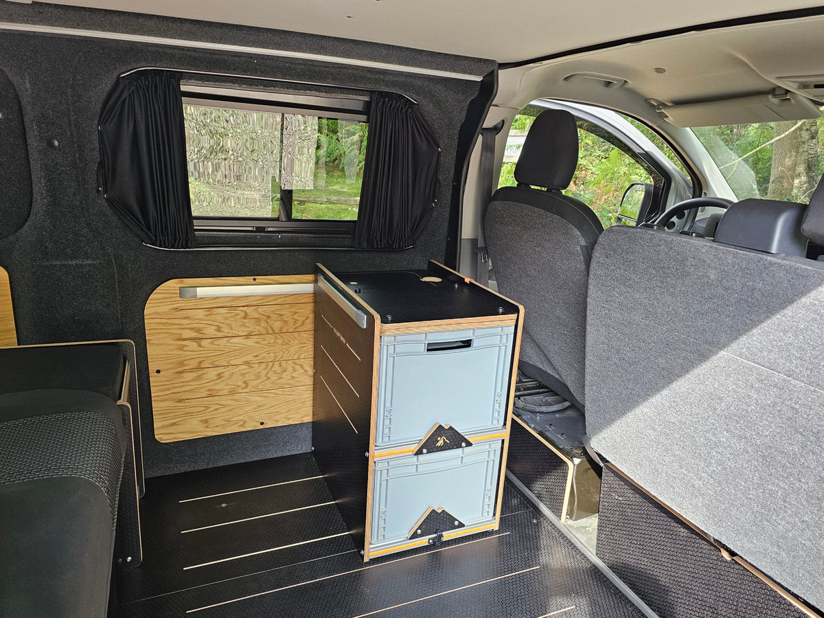 A compact camper storage cabinet with two light blue drawers and a black top, set inside a vehicle conversion setup with gray fabric seats and wood paneling.