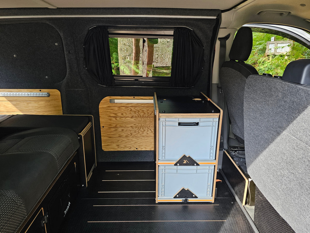 Custom camper van interior showing a light blue and wood storage unit, a black bench bed, and dark gray fabric seats.
