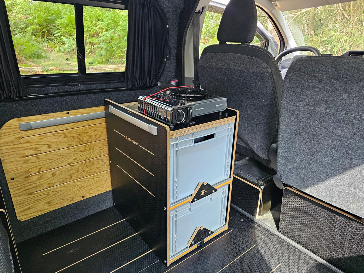 A compact black and wood storage cabinet with two light blue drawers sits in the back of a vehicle, with a portable camping stove on top. The interior features gray seats, a wood-paneled wall, and a window looking out onto green foliage.