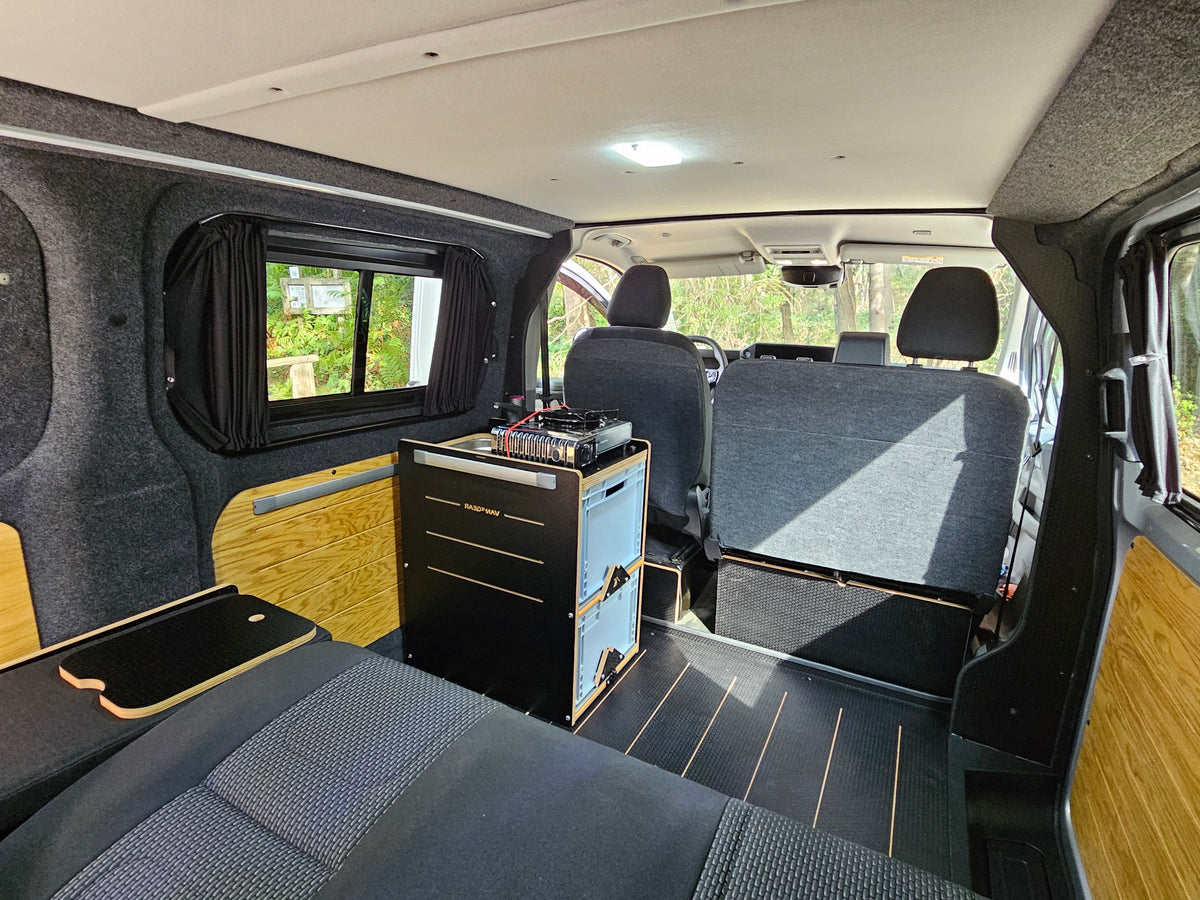 Rear cabin interior of a customized van with a raised black and blue utility storage unit, dark gray textured seats, wood-look paneling, and a window with dark curtains.