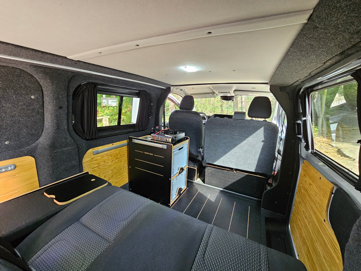 Rear interior of a converted van with a bed in the foreground, a small black and blue storage cabinet, wood-look paneling, and gray fabric seats.