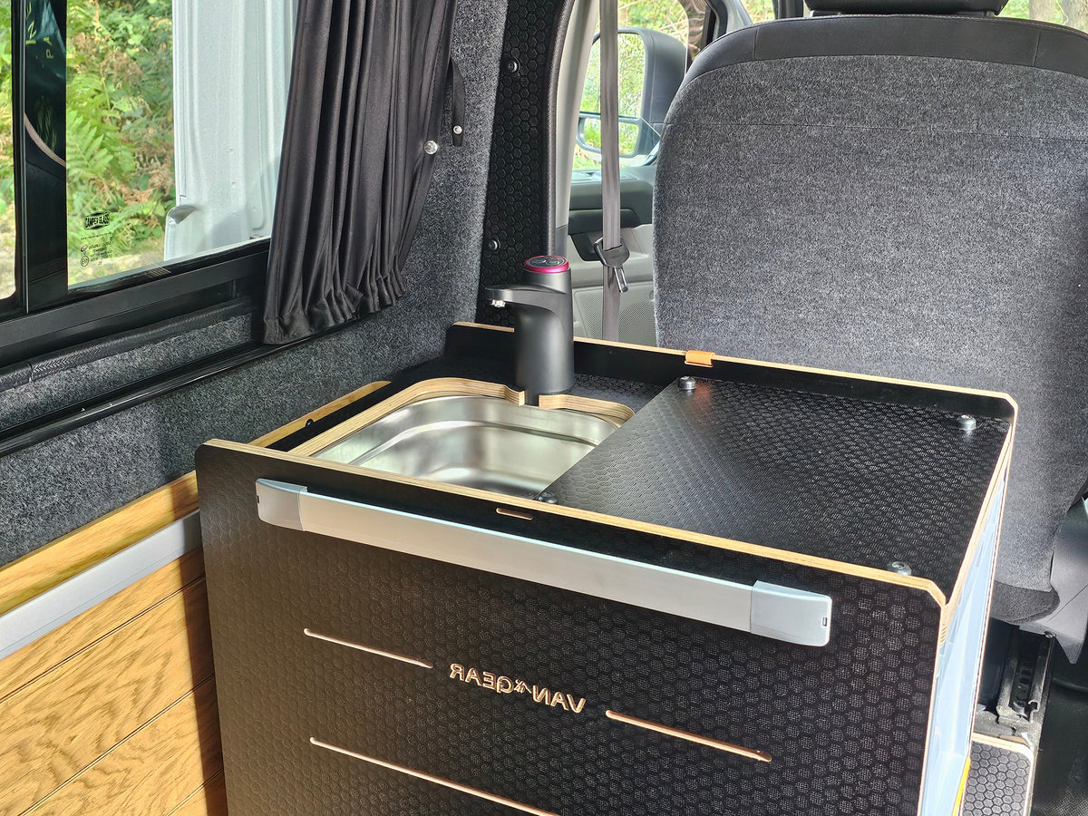 A compact black and wood vanity unit with an integrated stainless steel sink and dark faucet, mounted inside a vehicle or camper interior. The front panel shows the brand name VAN*GEAR.